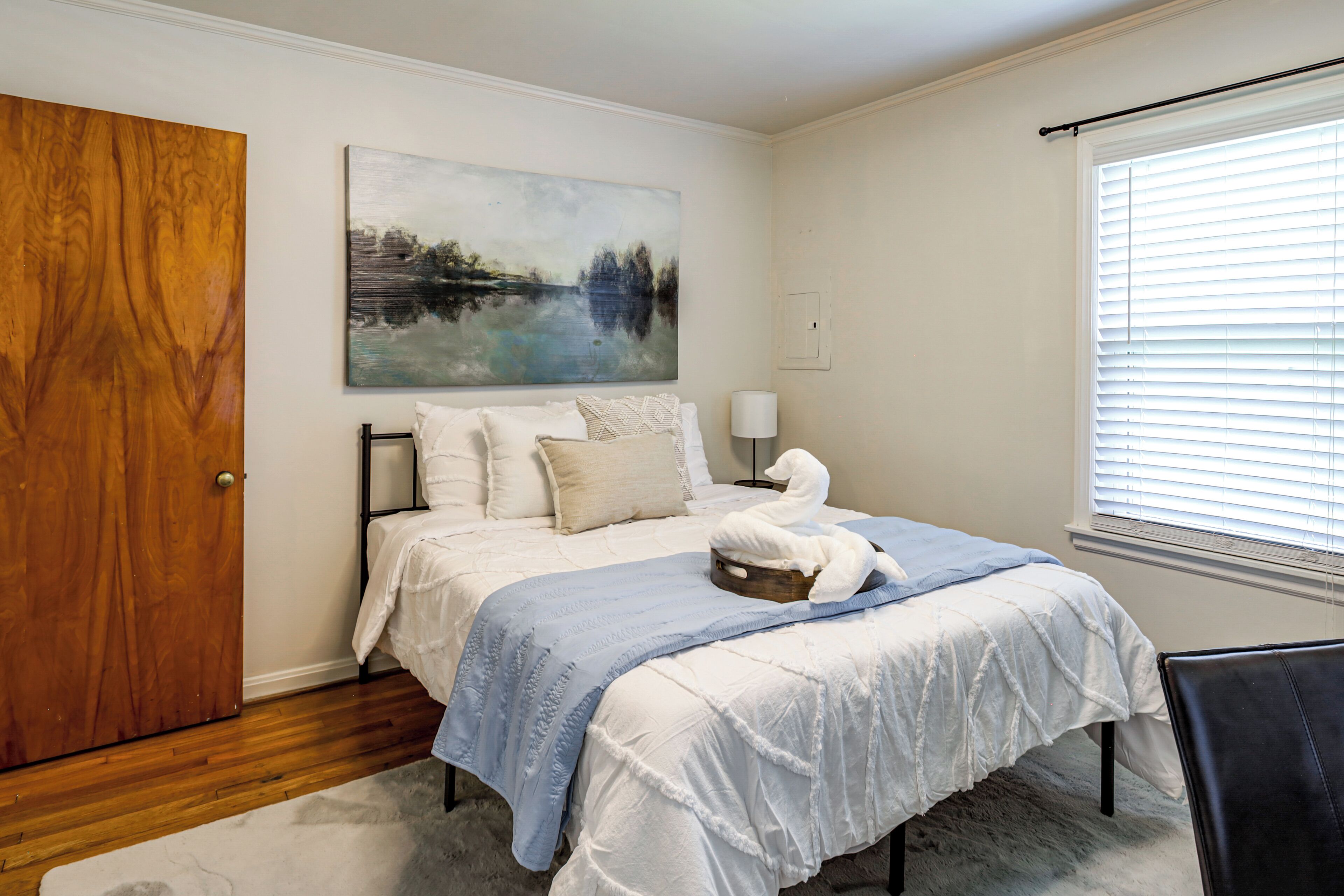 Un dormitorio con cama, puerta de madera, un cuadro, una ventana con persianas y una silla.