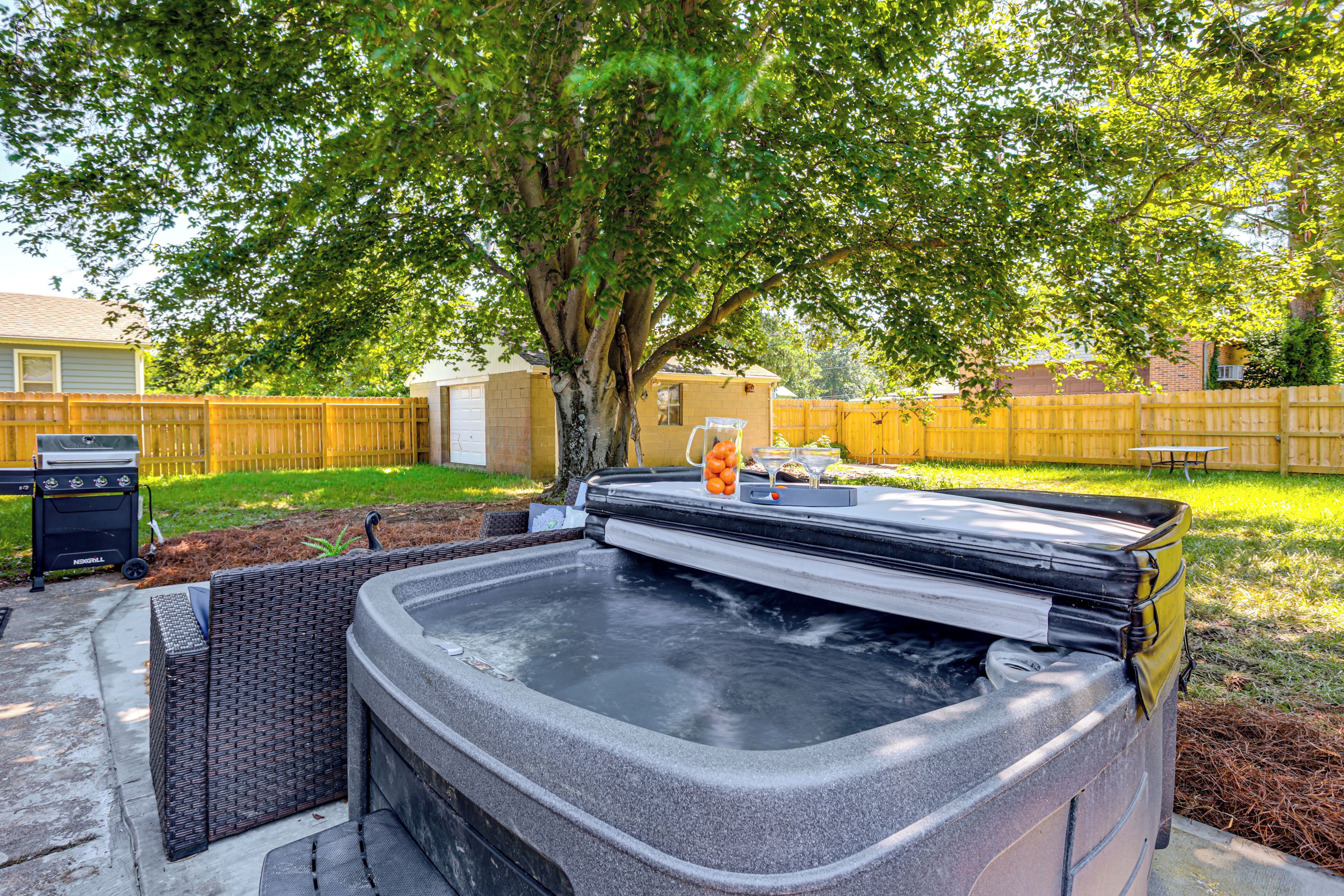 A hot tub in a backyard with a wooden fence, a tree, and a grill.