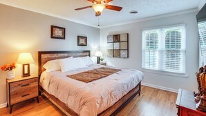 A bedroom with a wooden bed, a ceiling fan, a nightstand with a lamp, a window with blinds, and artwork on the wall.
