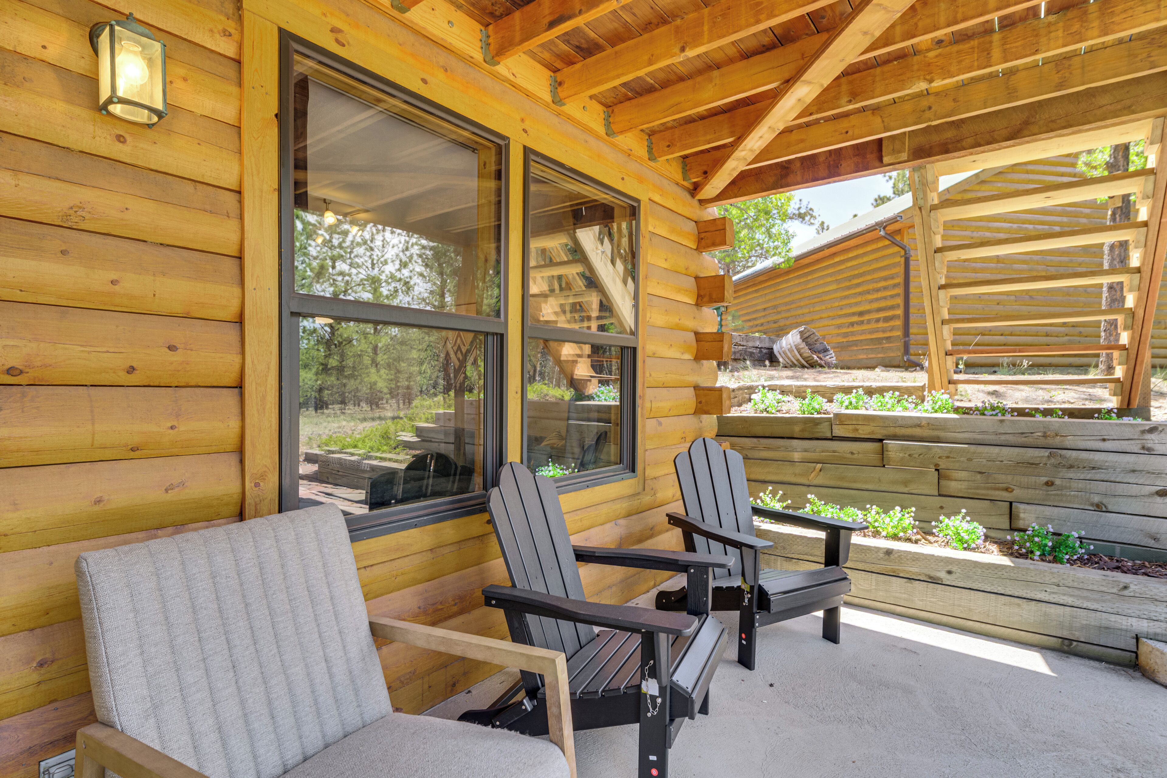 A log cabin with two Adirondack chairs, a wooden deck, and a large window overlooking a garden.