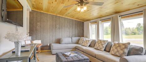 A living room with a sectional sofa, a grey ottoman, a ceiling fan, and a flat-screen TV mounted on the wall.