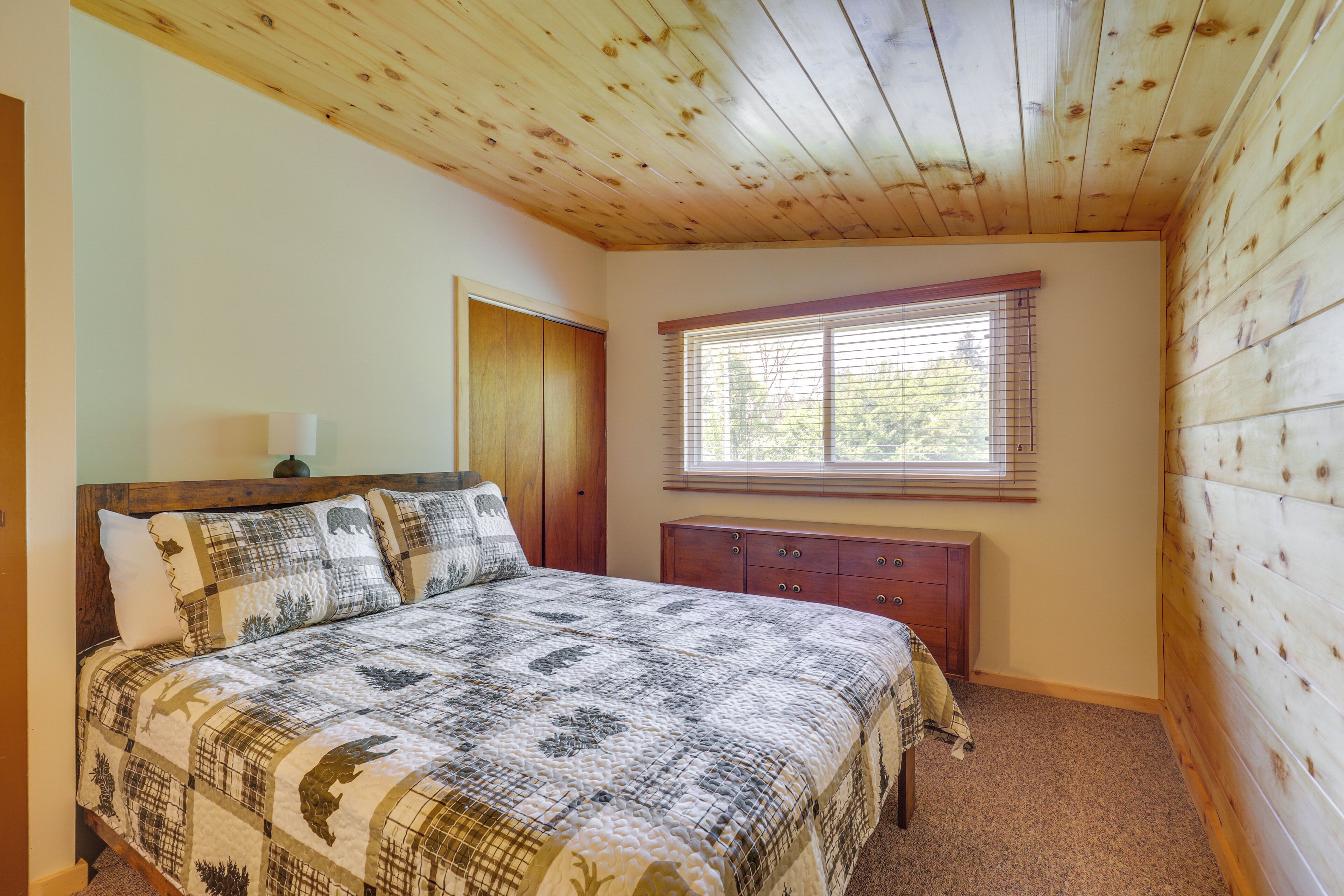 Een slaapkamer met een houten plafond, een bed met beddengoed met een patroon, een nachtkastje, een dressoir en een raam met jaloezieën.