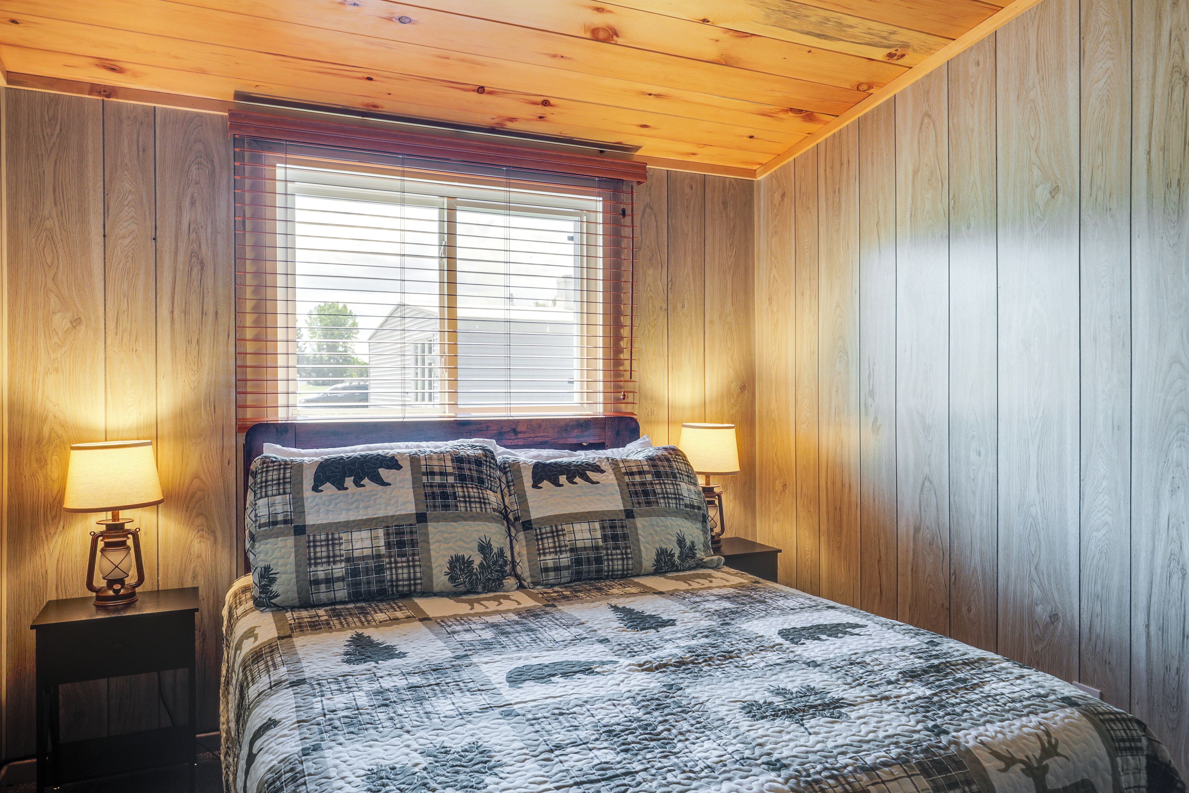 Quarto com teto de madeira, cama com colcha estampada, duas luminárias de cabeceira e janela com persianas.