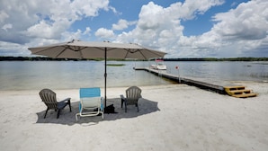 A beach with a table and chairs under an umbrella, a dock, and a boat in the background.