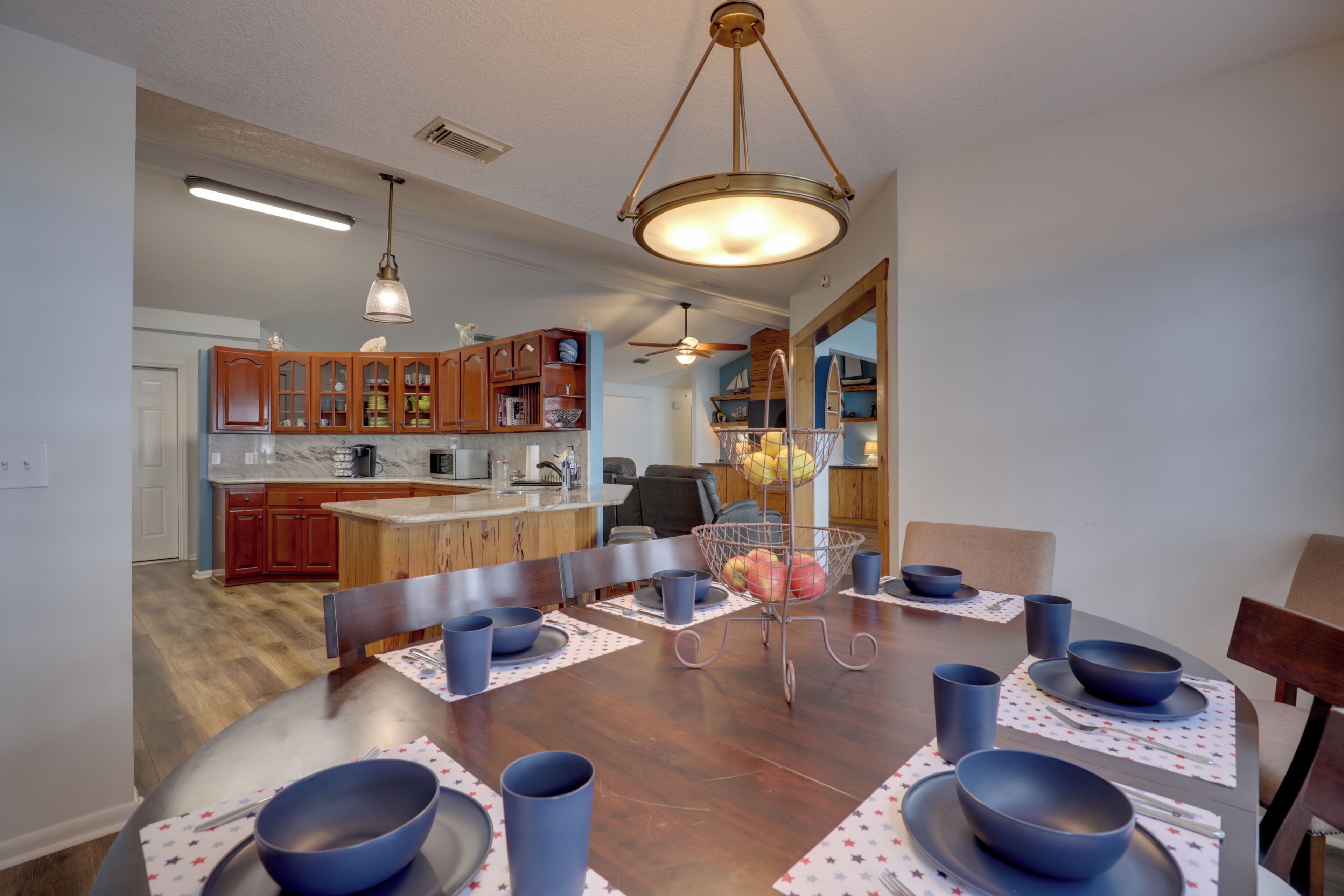 A dining area with a wooden table set for four, surrounded by chairs, with a kitchen in the background.