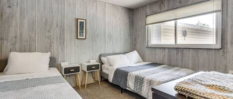 Une chambre avec deux lits, une fenêtre avec des stores, une table de chevet et un tableau accroché au mur.
