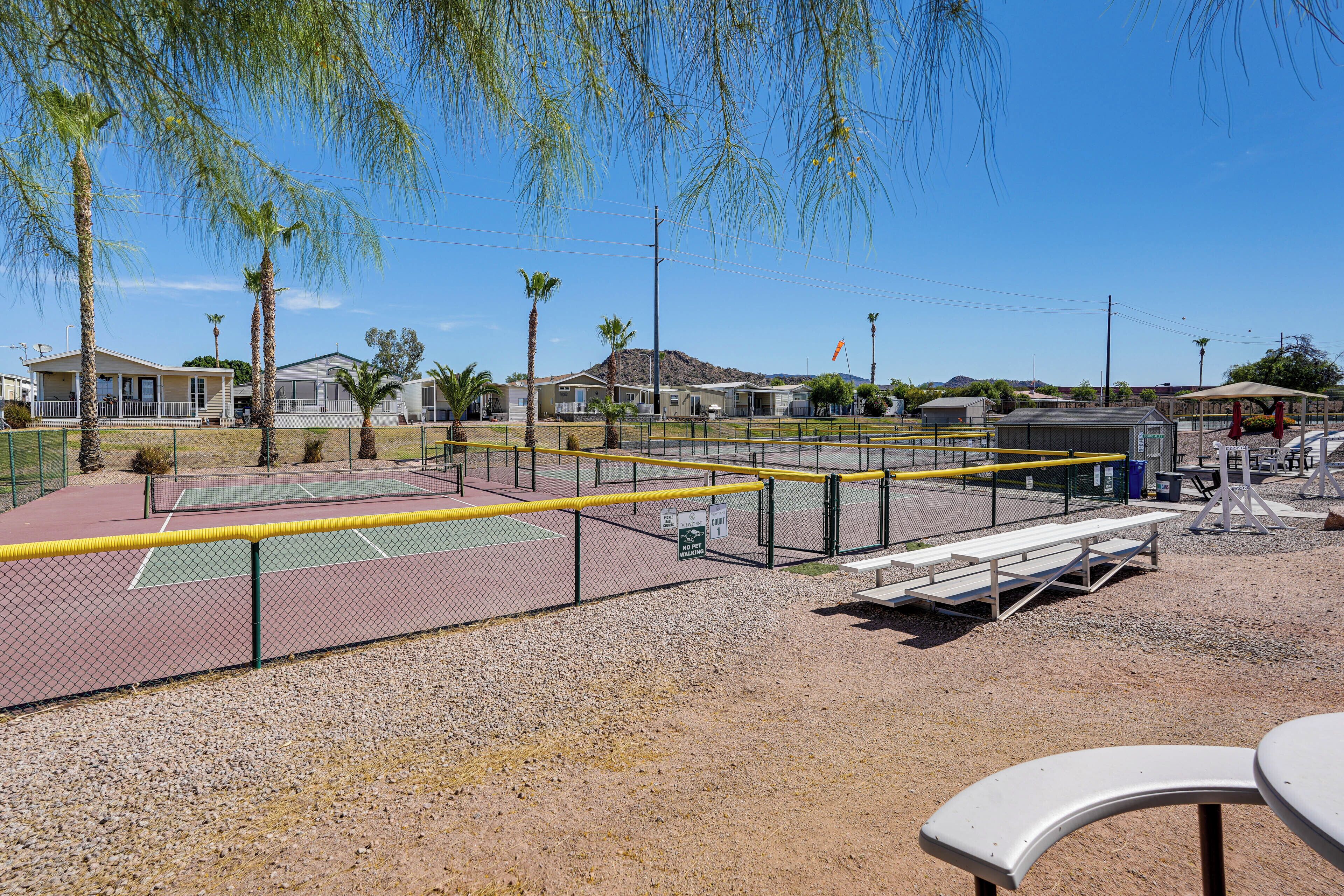 Un court de tennis entouré d’une clôture, avec des bancs et un petit bâtiment en arrière-plan.