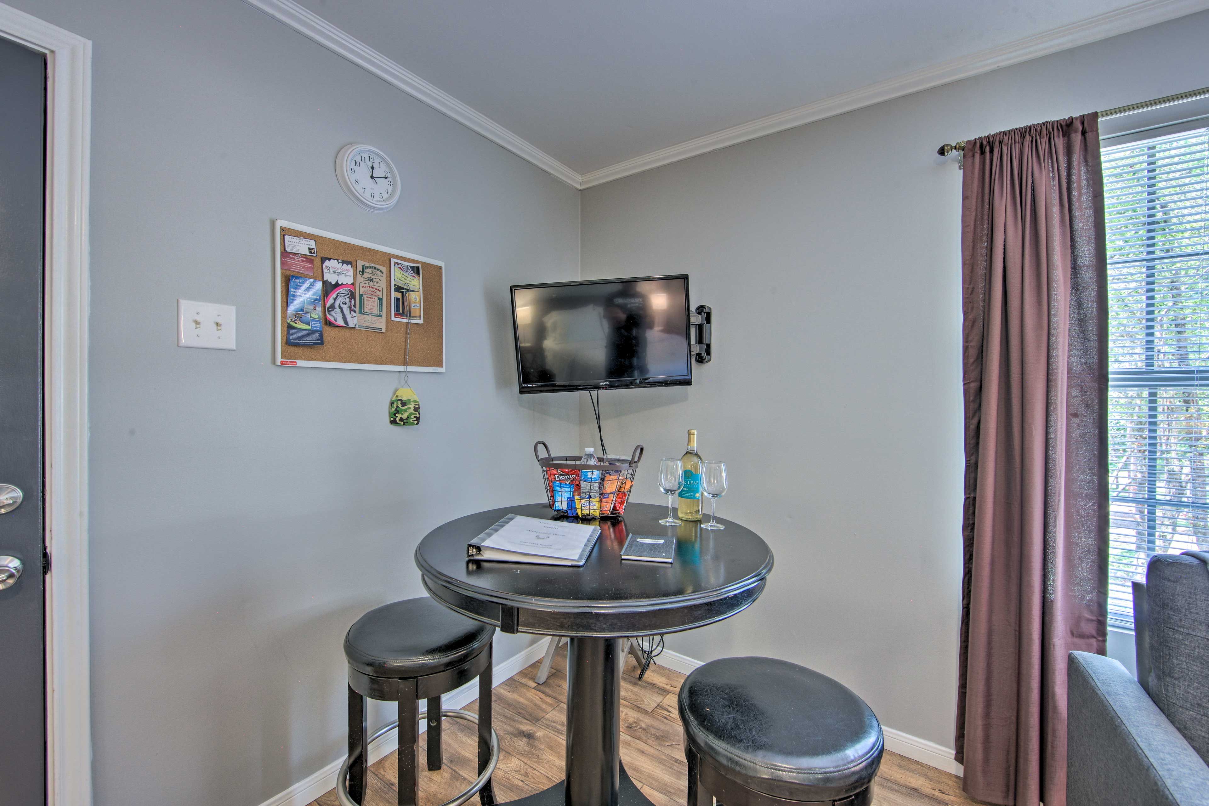 A small room with a round table, two stools, a TV mounted on the wall, a corkboard with notes, a window with curtains, and a door.