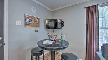 A small room with a round table, two stools, a TV mounted on the wall, a corkboard with notes, a window with curtains, and a door.