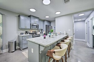 A modern kitchen with grey cabinets, a central island with bar stools, stainless steel appliances, and recessed ceiling lights.