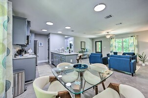 A modern living room with a glass dining table, blue sofas, and a kitchen area with grey cabinets and a central island.