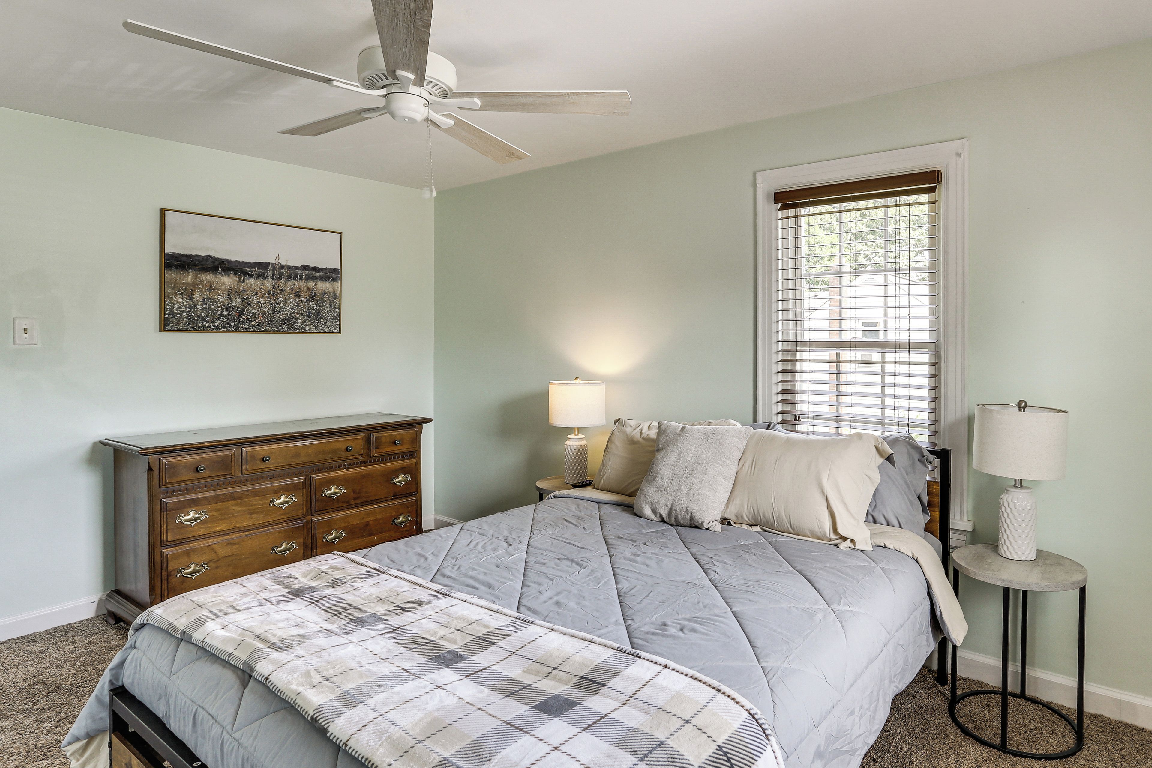 A bedroom with a bed, dresser, ceiling fan, window with blinds, lamp, and a framed picture on the wall.