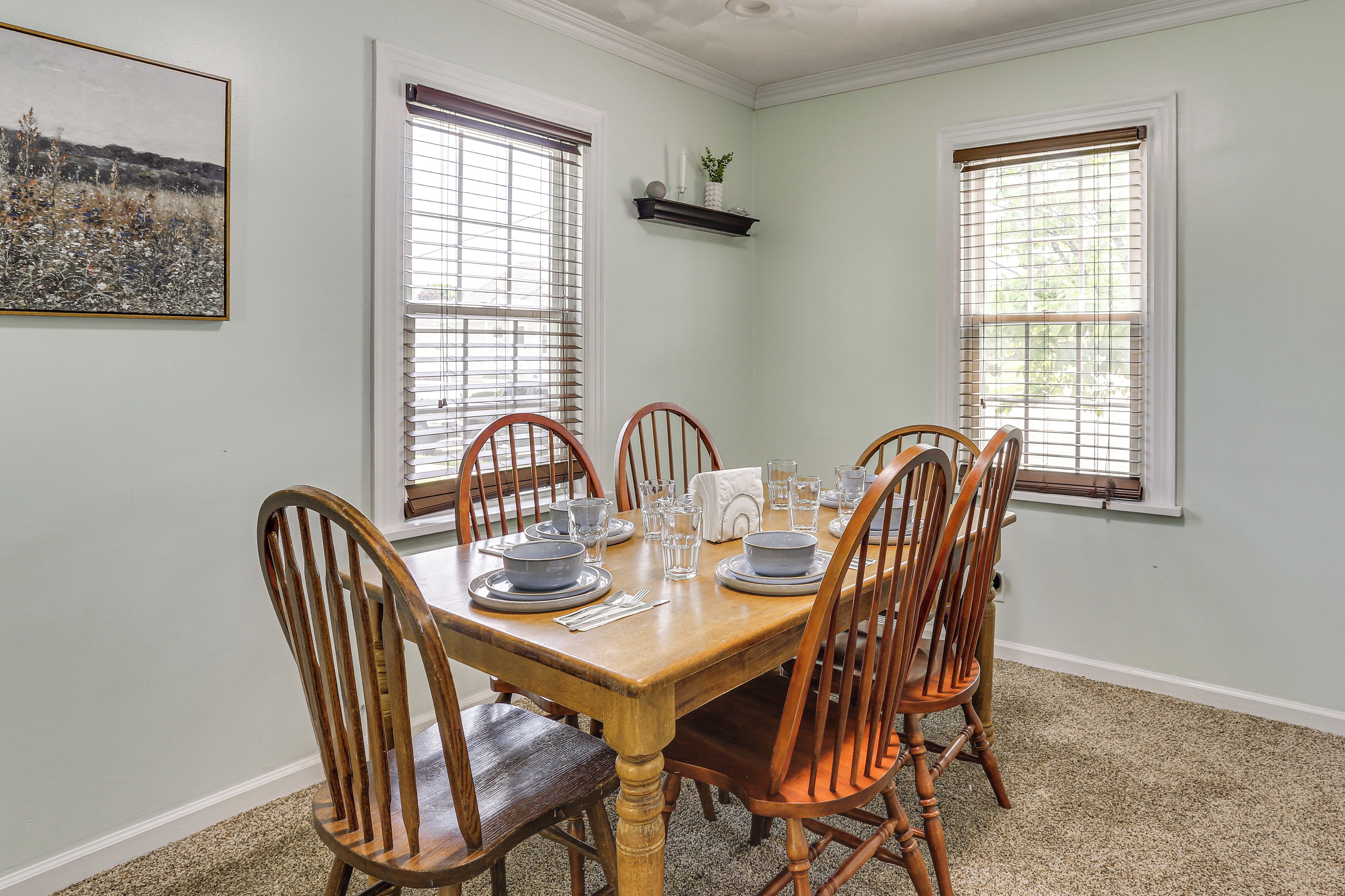 A dining room with a wooden table set for six, flanked by six wooden chairs, with two windows and a wall-mounted shelf above.