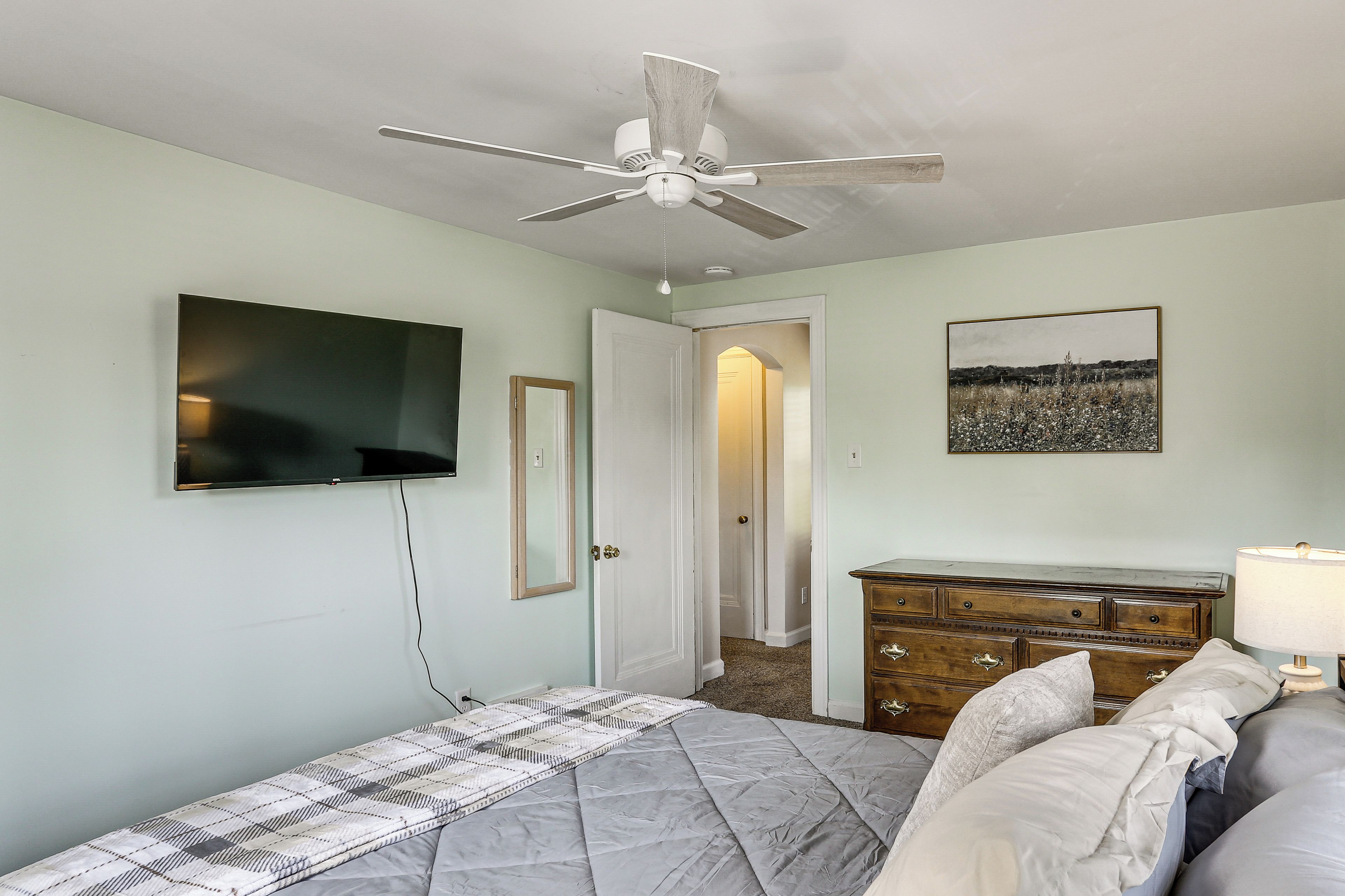 A bedroom with a bed, a ceiling fan, a dresser, a television, and a door leading to another room.