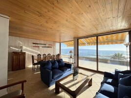 Amplia sala de estar con techo de madera, un sofá azul, comedor con sillas y vista al océano a través de amplios ventanales.