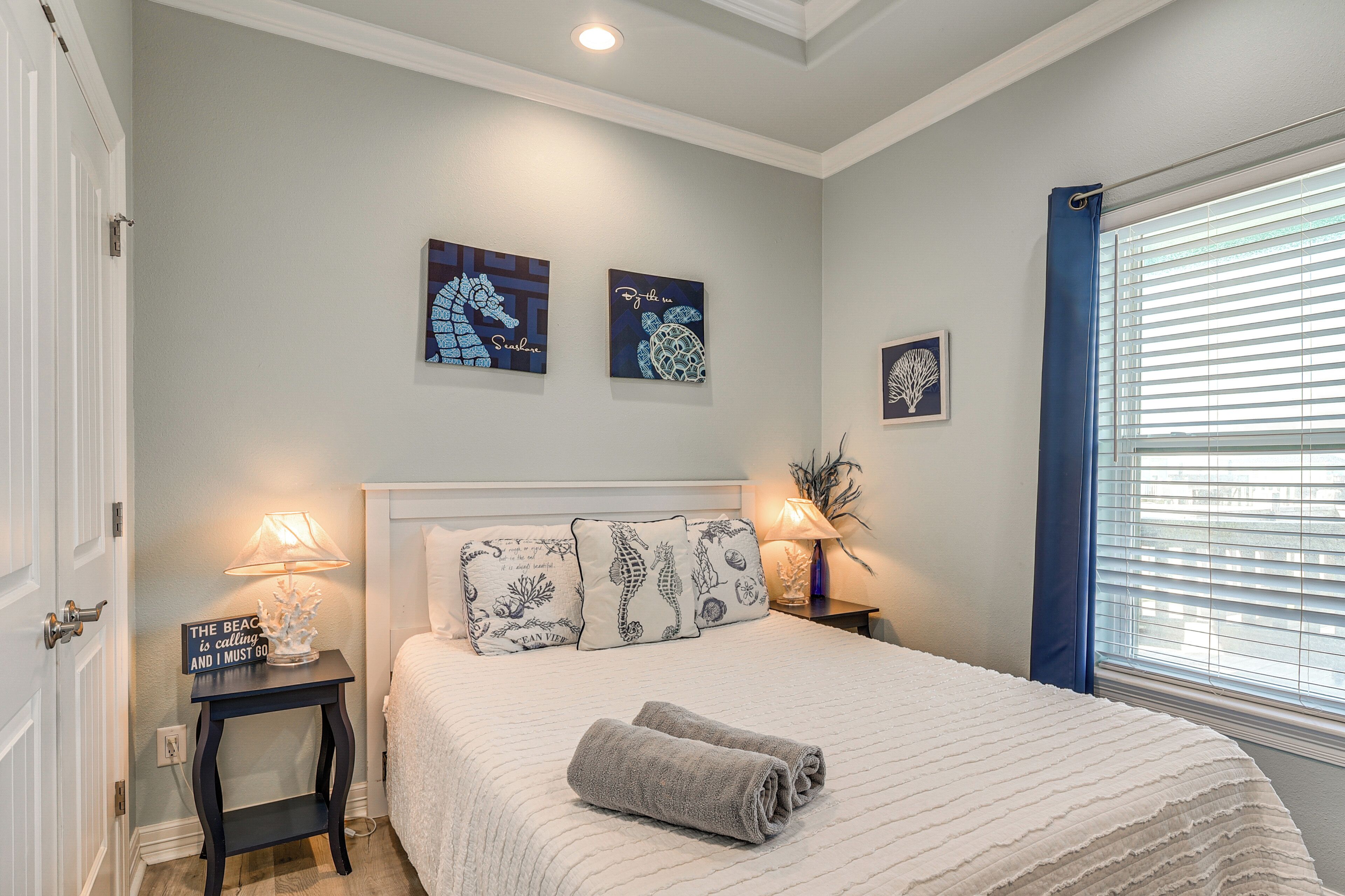A bedroom with a bed, two bedside tables with lamps, a wall with framed artwork, and a window with blinds.