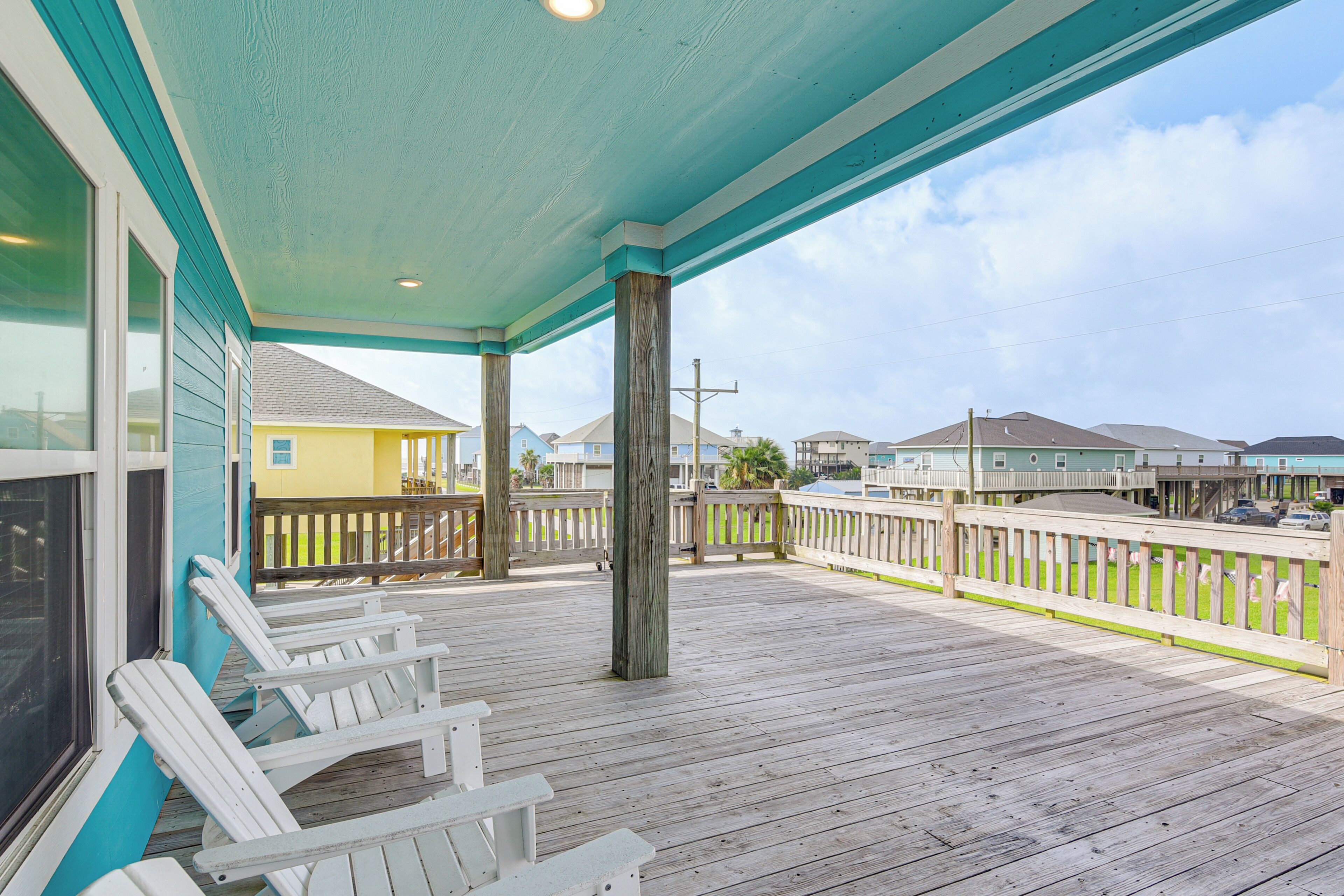 A wooden deck with white chairs, a covered area with teal trim, and a view of buildings in the background.
