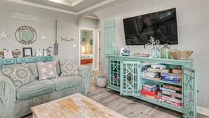 A living room with a teal sofa, a coffee table, a TV, and a cabinet with board games. - Bolivar Peninsula Beach House: Family Friendly! (Port Bolivar)