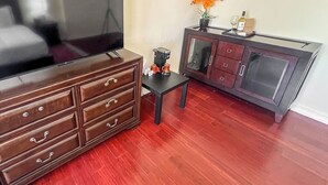 A room with a large flat-screen TV, a wooden cabinet with drawers, a small table with a coffee maker, and a vase with flowers.