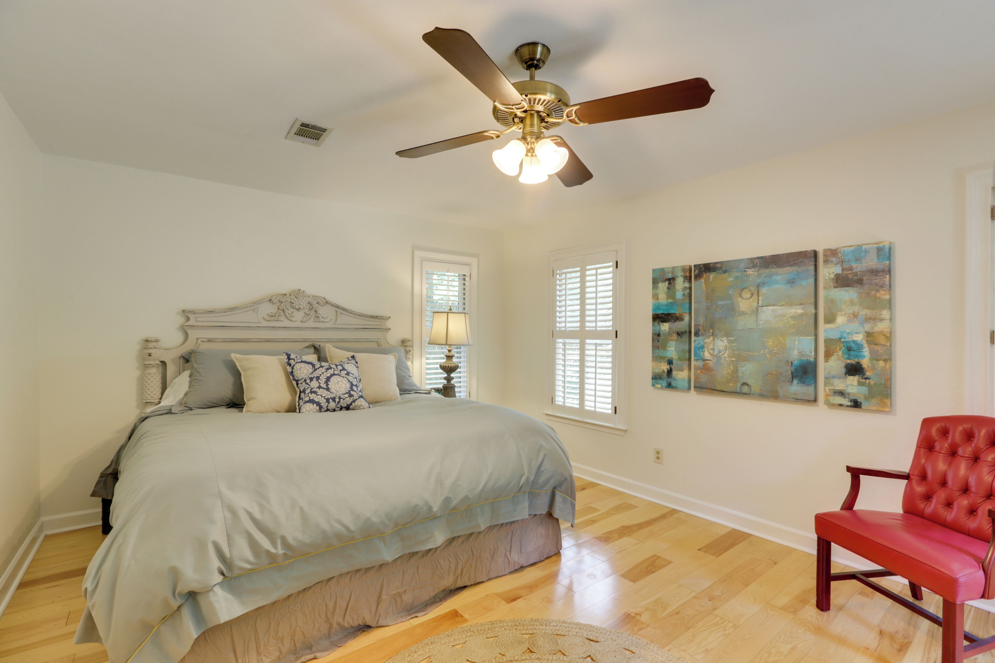 Une chambre à coucher avec un lit, une chaise rouge, un ventilateur de plafond et des œuvres d’art accrochées au mur.