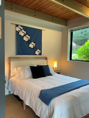 A bedroom with a bed, a nightstand, a wall-mounted lamp, a window with a view of greenery, and a blue wall hanging.