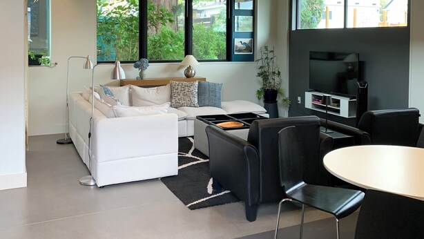 A modern living room with a sofa, a dining table, and chairs. There is a TV mounted on the wall and a window providing a view of the outdoors.