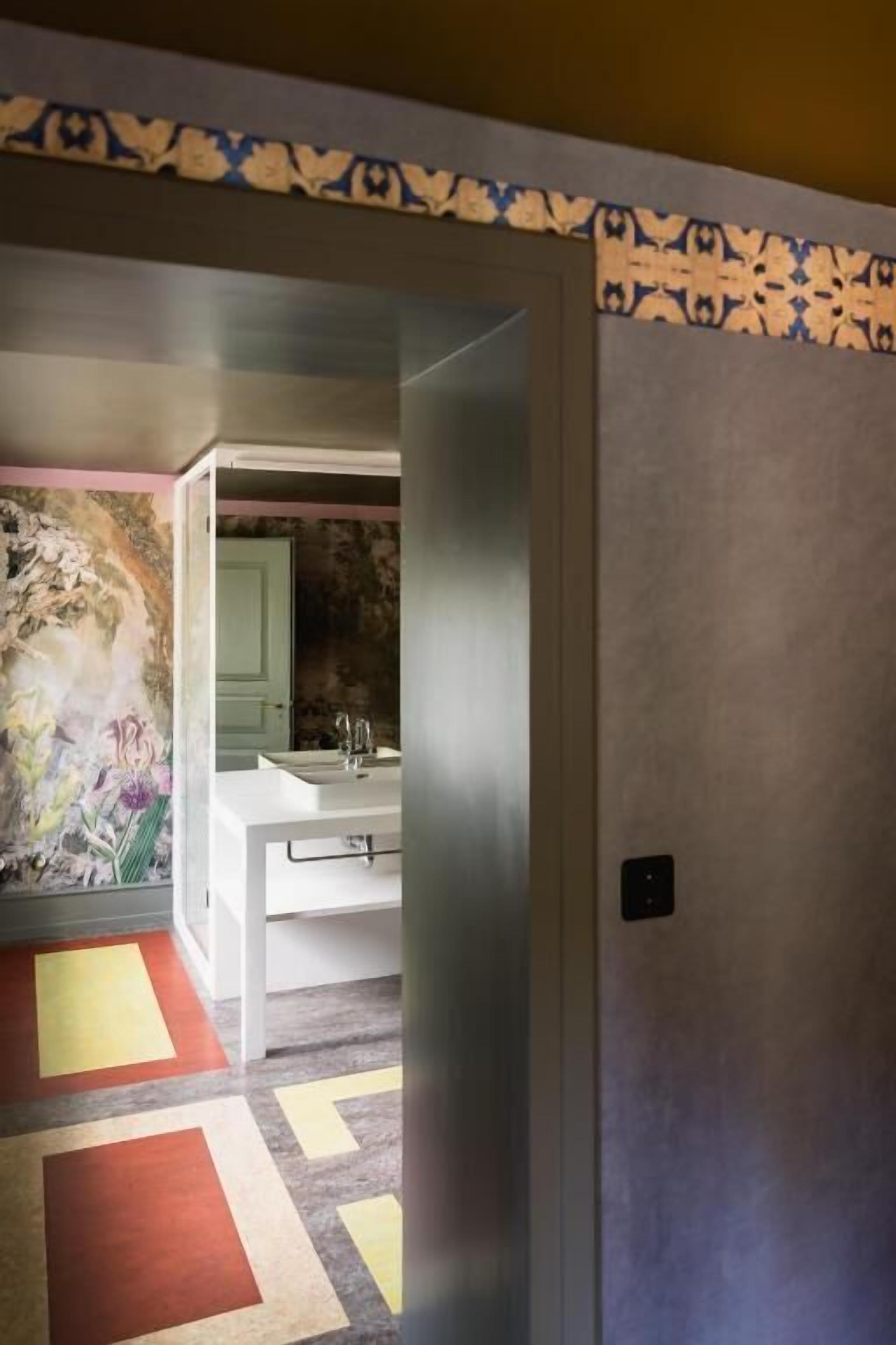 Un couloir orné d’une bordure à motifs, une salle de bain avec un lavabo et un miroir, et un sol aux couleurs vives.