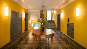 A hallway with yellow walls, patterned floor tiles, a round wooden table with a floral centerpiece, and a staircase in the background. - Post Hotel Löwe (Surses)