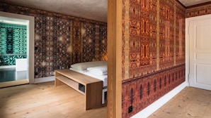 A bedroom with a wooden floor, a bed with white bedding, a bench, and a patterned wall. - Post Hotel Löwe (Surses)