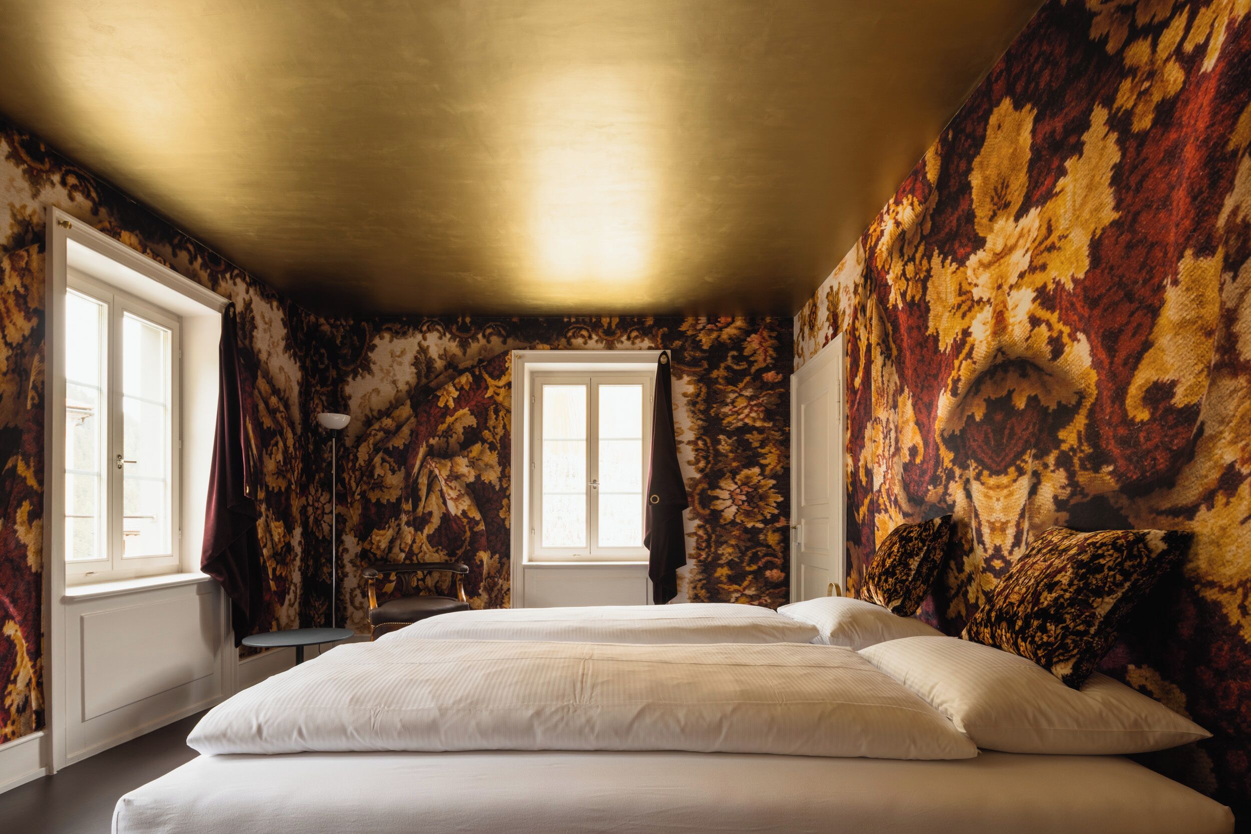 Un dormitorio con una cama grande, dos ventanas, una lámpara de pie y papel tapiz estampado.