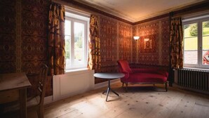 A room with patterned wallpaper, a red sofa, a small round table, and a window with curtains. - Post Hotel Löwe (Surses)