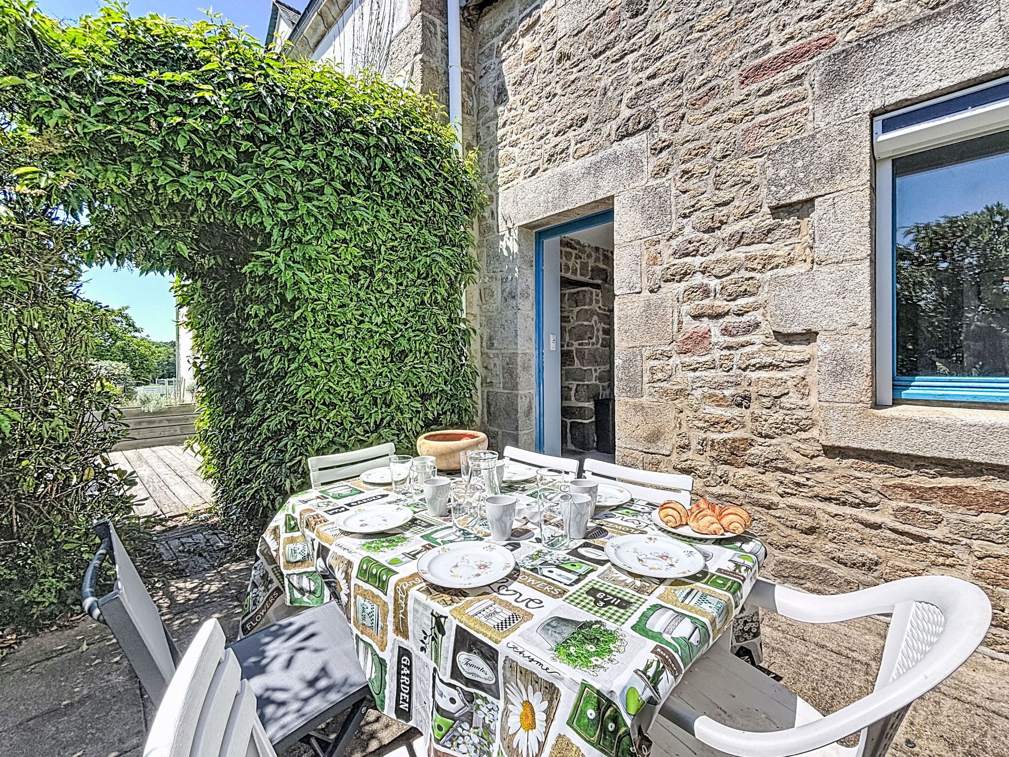 A table set outdoors with a patterned tablecloth, surrounded by stone walls and a leafy hedge.