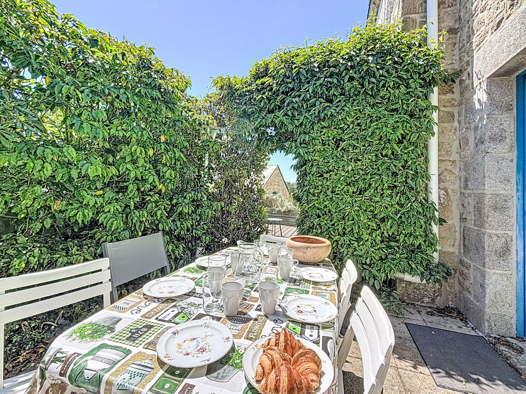 An outdoor dining area with a table set for two, surrounded by lush greenery and stone walls.