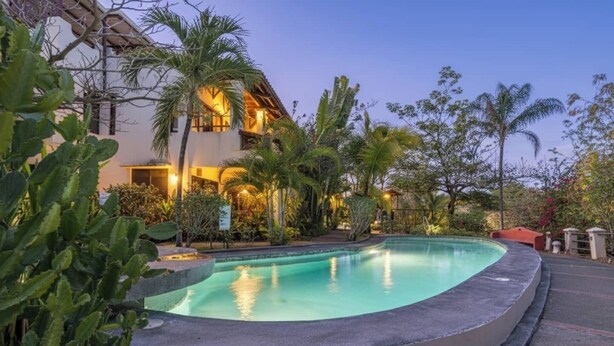 Une piscine à bords incurvés, entourée d’une végétation tropicale et bordée d’un bâtiment à l’éclairage chaleureux.