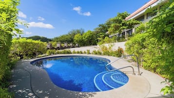 A swimming pool with a curved edge, surrounded by greenery and a building with a red roof.