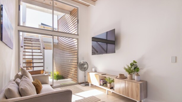 A modern living room with a sofa, a TV mounted on the wall, a wooden console table with decorative items, and a sliding glass door leading to an outdoor area. - Modern Pool Home in Coco · Walk to Beach and Town (Playa del Coco)