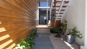 A modern building with wooden siding, a glass door entrance, and a small garden area with plants and a gravel path. - Modern Pool Home in Coco · Walk to Beach and Town (Playa del Coco)