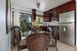 A kitchen with wooden cabinets, a stainless steel refrigerator, a glass dining table with wicker chairs, and a view of the outdoors through a sliding glass door. - Beach Escape in Las Palmas•Pool•Walk to Everything (Playa del Coco)