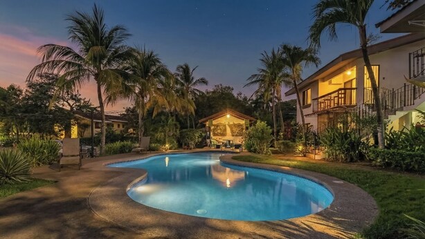 Uma piscina com borda curva, rodeada por palmeiras e um edifício com varanda.