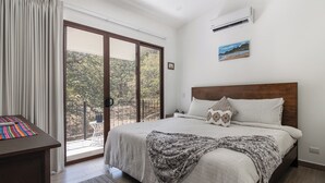 Et soveværelse med seng, skrivebord, stol, balkon med udsigt til træer og en aircondition-enhed på væggen.