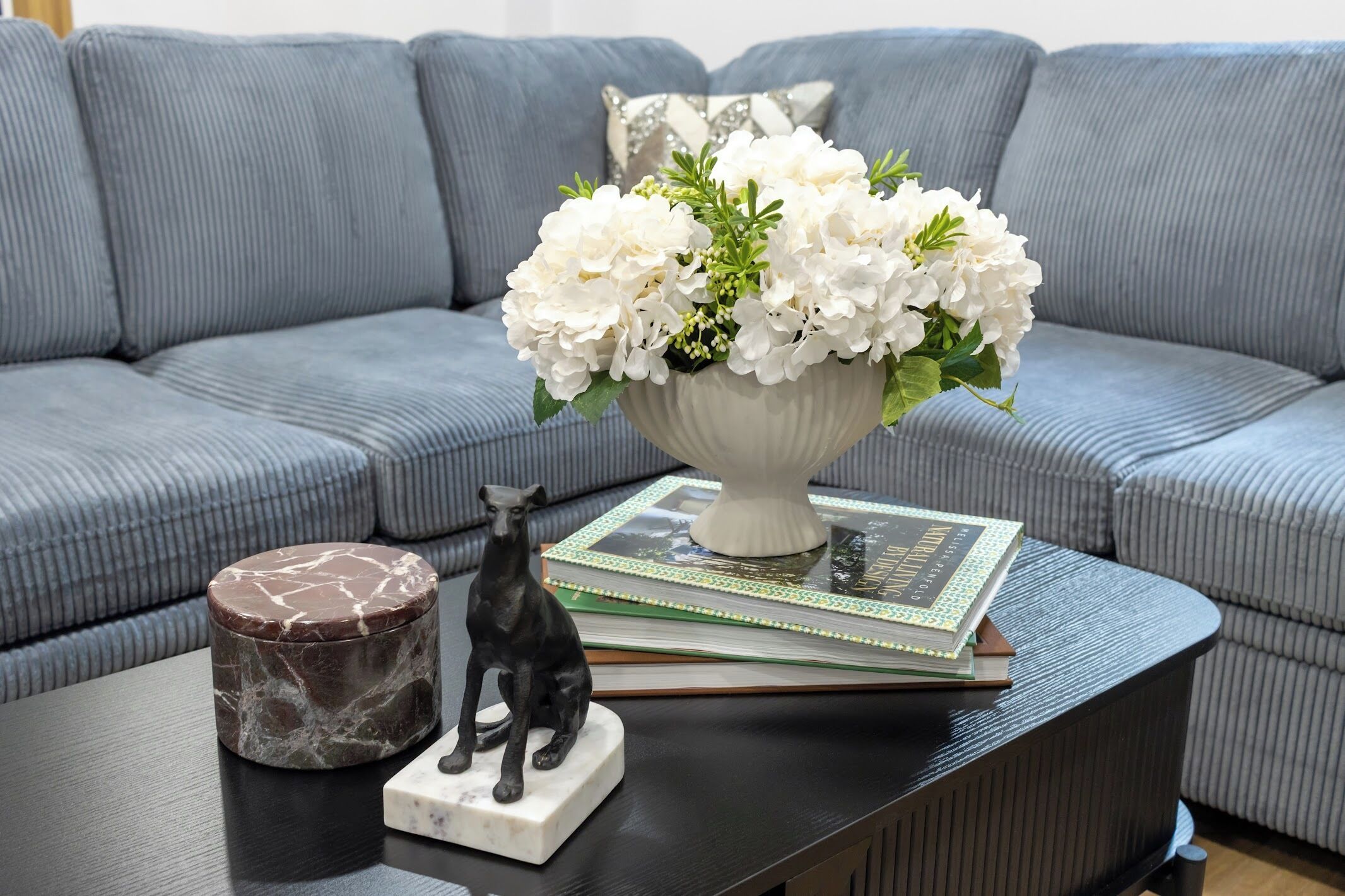 A grey sofa, a white vase with flowers, a book, a marble container, and a black statue on a black table.