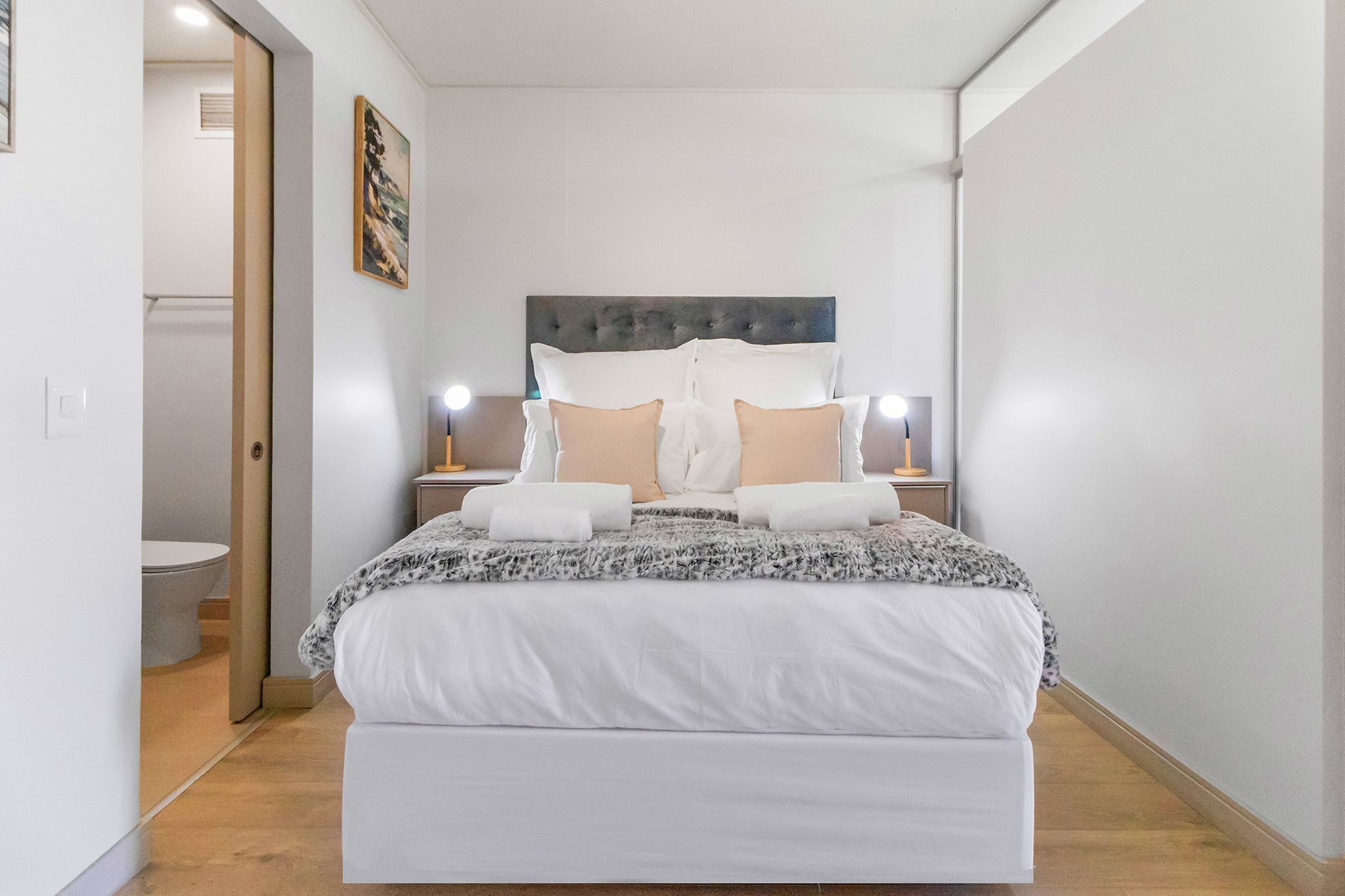 A modern hotel room with a large bed, bedside lamps, a headboard, and a painting on the wall.