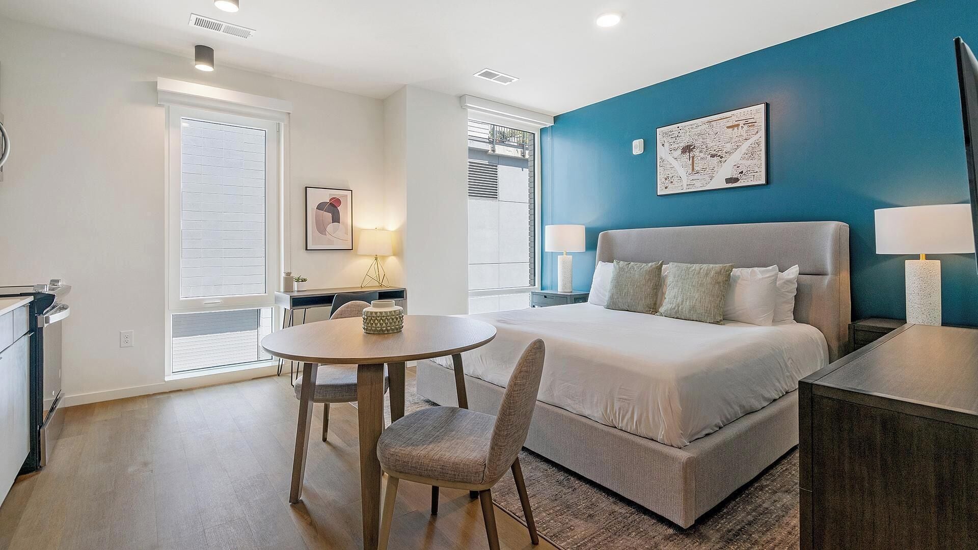 A modern bedroom with a large bed, a bedside table, a chair, a round table, and a window with blinds.