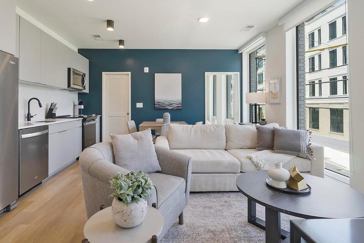 A modern living room with a grey sectional sofa, a small round table with a plant, and a kitchen area with stainless steel appliances.
