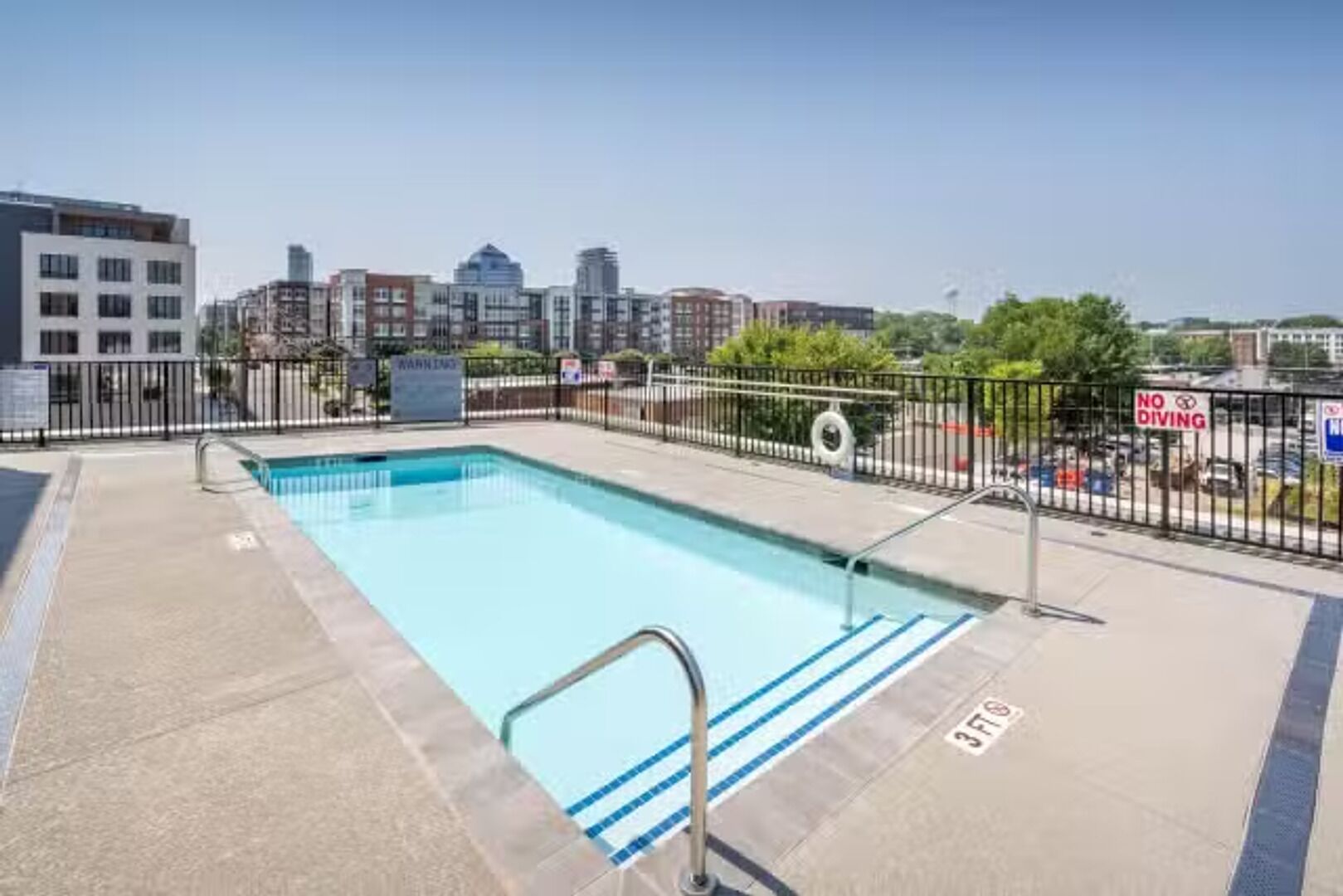 A rooftop pool with a city view, surrounded by a metal fence, and a 'No Diving' sign.
