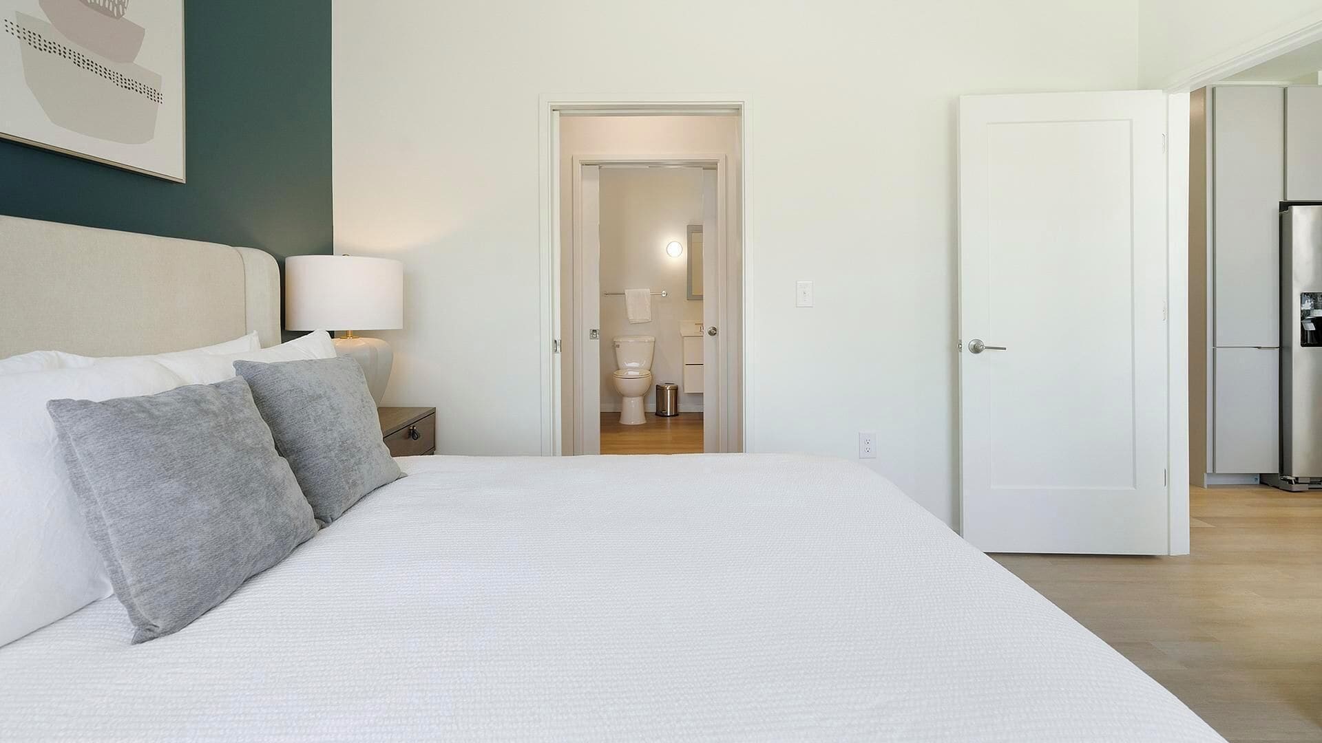 A bedroom with a large bed, a nightstand, a lamp, and a door leading to a bathroom.