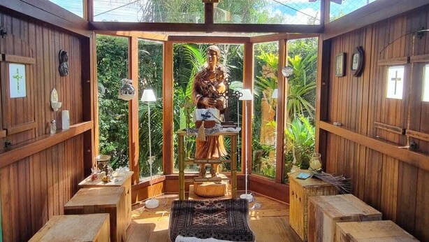 A wooden interior with a central statue, wooden benches, and a glass enclosure with greenery visible outside. - Casarão Hortência (Teresópolis)
