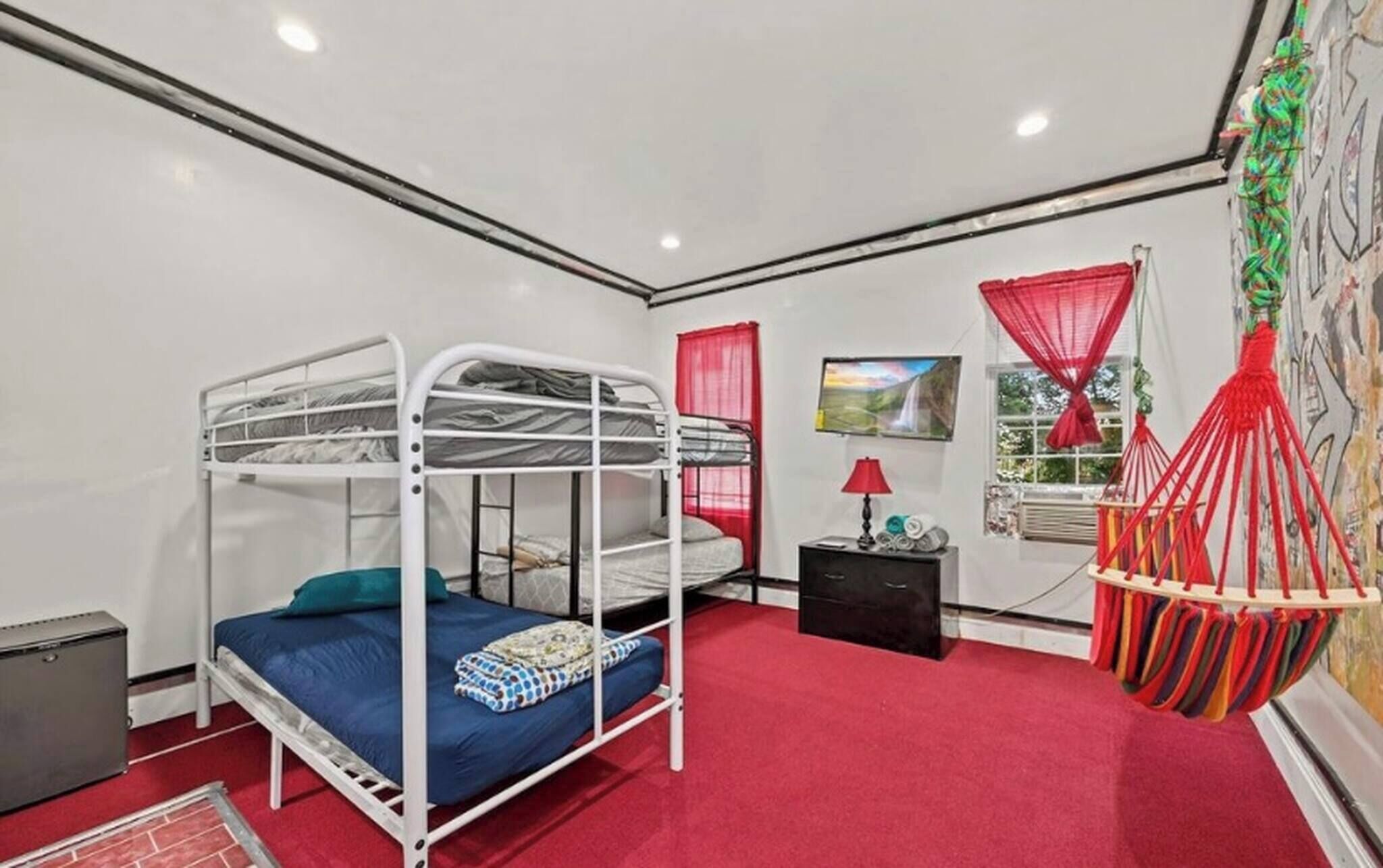 A bunk bed room with a red carpet, a small desk with a lamp, a window with red curtains, and a hanging chair.