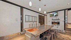 A modern kitchen with a central island, bar stools, pendant lighting, a fireplace, and a TV mounted on the wall.