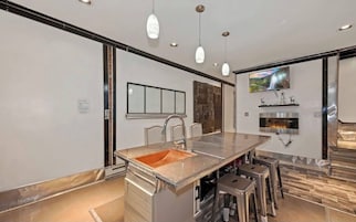 Una cocina moderna con una isla central, taburetes de bar, iluminación colgante, chimenea y un televisor montado en la pared.